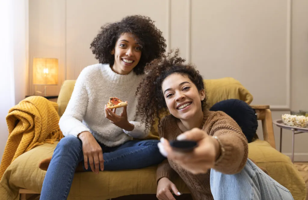 Two young women watching TV and smiling while eating pizza, representing modern tv viewership statistics trends.